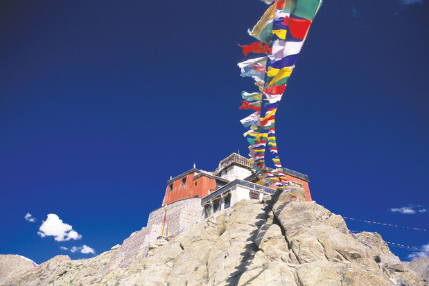 A multicolored string of flags flutters above a rugged rocky path leading to a red-and-white building, set against a clear blue sky with scattered clouds.